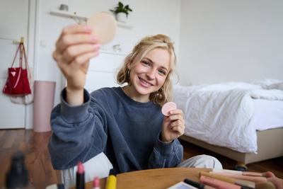 Image of young blond woman, girl records video about makeup, shows skin tone beauty products, sits in a room on floor Im-stock-foto