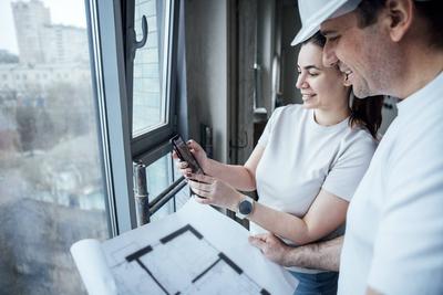 Ein l?chelnder Mann mit Helm schaut sich sorgf?ltig einen Bauplan an und hilft bei der Planung der Arbeiten. Eine Frau h?lt ein Telefon in der Hand und zeigt Dekor- und Oberfl?chenelemente.-stock-foto