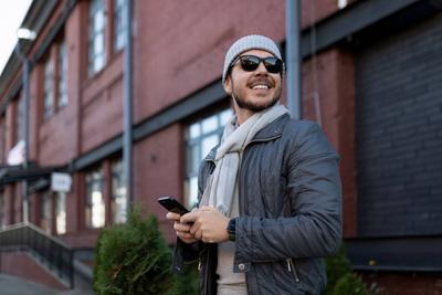 starker junger Mann mit Sonnenbrille, der ein Telefon in der herbstlichen Stadt h?lt-stock-foto