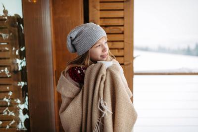 Smilng teen girl bundled in thick blanket holding cup of hot tea standing on balcony during winter day.-stock-foto